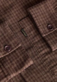 Chemise en tissu brun texturé avec deux poches poitrine boutonnées et une petite étiquette de marque noire et dorée entre elles.