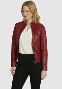 Rote Lederjacke mit Reißverschluss vorn, Stehkragen und taillierter Passform. Kombiniert mit einer weißen Bluse und schwarzen Hosen mit Reißverschlusstaschen.