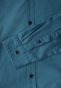 Tyrkysová košile s dlouhým rukávem a manžetou na knoflíky, s černými knoflíky a hladkou texturou, úhledně složená pro vystavení.