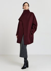 Burgunderfarbener, oversized Wollmantel mit hohem Kragen und asymmetrischer Front. Tragend über einem grauen, figurbetonten Rock und schwarzen, knielangen, hochhackigen Stiefeln.