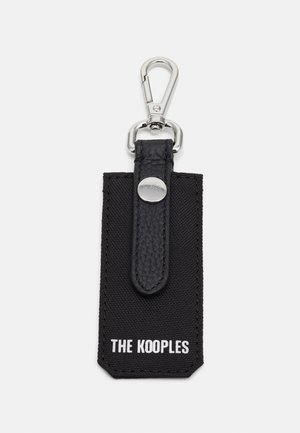 Melnas atslēgu piekariņš ar teksturētu ādas siksniņu, metāla sprādzi un taisnstūra auduma birku, uz kuras balta krāsā ir izdrukāts "THE KOOPLES".