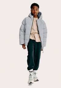 Lichtgrijze pufferjack met capuchon, doorgestikt ontwerp, ritssluiting, gecombineerd met donkere trainingsbroek en lichtgekleurde sneakers.