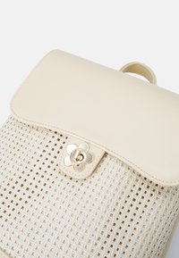Borsa di colore crema con una texture intrecciata, chiusura floreale dorata e patta in pelle liscia. Design unico che combina materiali e finiture.