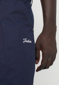 Pantaloni bleumarin dintr-un material texturat, cu un logo alb brodat "Tacker" și accente cu nasturi pe posetele laterale.