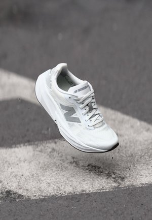 Biały but do biegania New Balance w powietrzu nad asfaltem z białymi oznaczeniami drogowymi i małymi odpadkami.