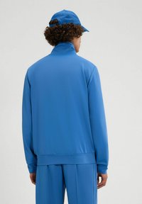 Sudadera azul con cuello alto, textura suave, mangas largas y cinturilla acanalada. Combinada con una gorra azul a juego.