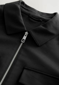 Chemise zippée noire avec un col, en tissu lisse. Dispose d'une fermeture éclair argentée et d'une poche poitrine. Lignes épurées et design moderne.