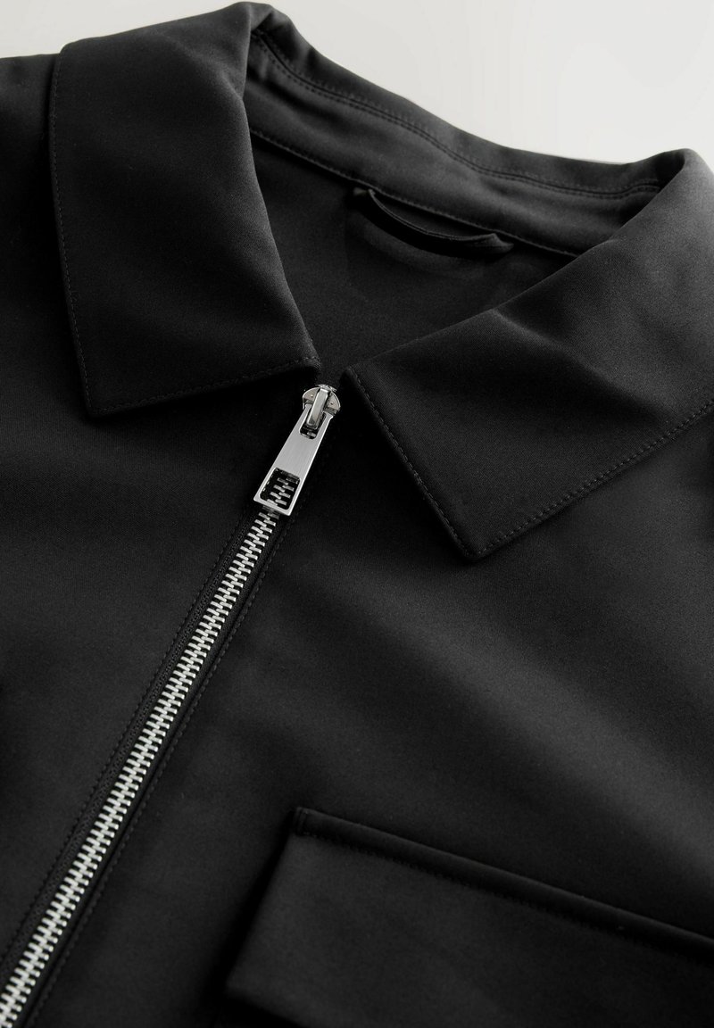 Chemise zippée noire avec un col, en tissu lisse. Dispose d'une fermeture éclair argentée et d'une poche poitrine. Lignes épurées et design moderne.