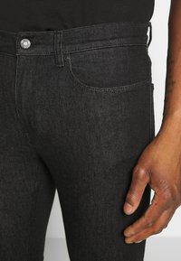 Černé džíny z denimu mají pět kapes, kontrastní prošívání a kovové zapínání. Materiál má hladkou texturu s mírným leskem.