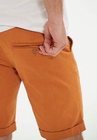 Kurze Hosen in einem warmen Orangeton mit glatter Textur; verfügen über eine Gesäßtasche mit Knopfverschluss und gefaltete Saumdetails.