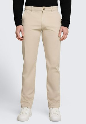 Homme portant un pantalon beige droit, des baskets blanches et un haut noir à manches longues, debout, les mains détendues le long du corps.
