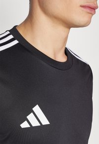 Black athletic t-shirt made of smooth fabric. Features white three-stripe accents on shoulders and a large white logo on the chest.