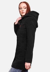 Black hooded coat with a textured fabric, zip closure, side pockets, and a logo detail near the hem, styled with light blue jeans.