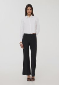 Camicia bianca a maniche lunghe con colletto e bottoni, abbinata a pantaloni neri svasati e sandali neri con fasce. Tessuto liscio, design minimalista.