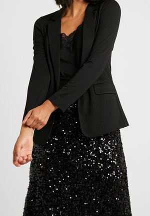 Veste noire élégante avec un design épuré, dotée de revers crantés et de deux poches avant. Portée sur un caraco en dentelle noire et une jupe à sequins.