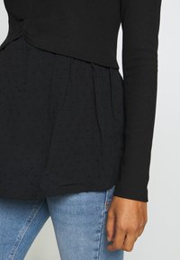 Rechter Arm und Taille einer Person, die eine schwarze strukturierte Bluse über hellblauen Jeans mit sichtbarem Taschen-Detail trägt.