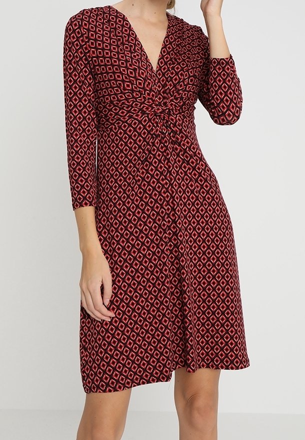 Femme portant une robe rouge mi-longue avec des motifs géométriques en losange roses et des manches trois-quarts, posant avec une main près de son visage.