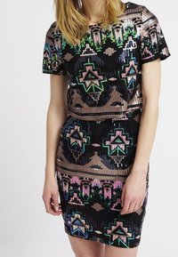 Černé šaty s krátkými flitrovými rukávy a složitým geometrickým vzorem v zelené, modré, růžové a bronzové barvě. Upnutý střih zvýrazňuje siluetu.