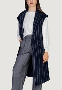 Námořnický modrý dlouhý vest s bílými vertikálními pruhy, velkým límcem, knoflíky a bočními kapsami; nošený přes bílý svetr s kabelovým vzorem a šedé kalhoty.