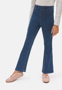 Jeans de denim azul acampanados con una cintura alta, que presentan costuras visibles y bolsillos laterales. Combinados con zapatos de tela rosa claro.