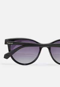 Ochelari de soare tip cat-eye cu rame din plastic negru, lentile violet cu un gradient și logo-ul aurit "Polaroid" pe brate. Design elegant și geometric.