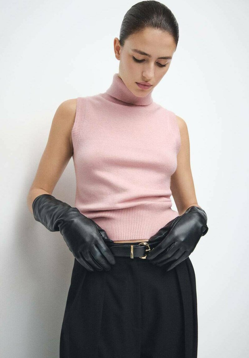 Crne kožne rukavice na osobi koja nosi svijetlo ružičasti top bez rukava s visokim ovratnikom i tamne hlače, ističući crni remen sa zlatnom kopčom.