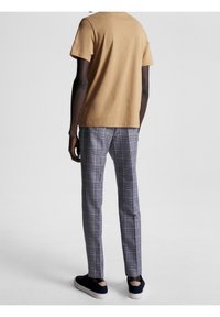 Man med beige kortärmad skjorta, grå rutig byxor och svarta sneakers, stående med ryggen mot kameran på en enkel bakgrund.