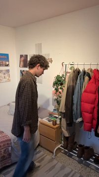 Jeune personne en chemise à carreaux se tient dans la chambre à côté du lit et d'un portant avec des vestes et des bottes, près d'une table de chevet en bois avec des plantes.