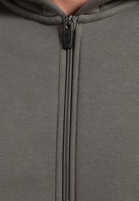 Grå lynlås hoodie med en glat tekstur, der har en metal lynlås og hætte. Stoffet er strækbart og designet til komfort og bevægelsesfrihed.
