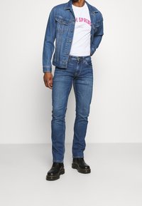 Jeansjacka i ljusblått, med framfickor och klassisk sömnad. Buren över en vit t-shirt med rosa tryck och mörka jeans. Svarta stövlar.