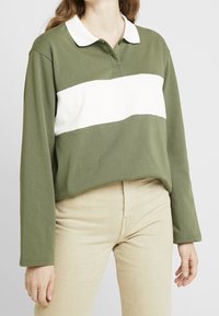 Femme portant un polo à manches longues olive et blanc avec un pantalon beige, debout devant un fond clair uni.