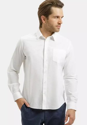 Mężczyzna o jasnobrązowych włosach, ubrany w białą koszulę zapinaną na guziki z kieszenią na piersi oraz ciemnoniebieskie spodnie, patrzący w prawo.