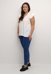 Weiße, kurzärmlige Bluse mit Rüschenschultern, kombiniert mit eng anliegenden blauen Jeans und schwarzen Loafers, stehend vor neutralem Hintergrund.