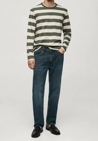 Mann trägt ein grün-weiß gestreiftes langärmeliges Hemd, blaue Jeans, schwarze Schuhe und steht mit einer Hand in der Tasche vor einem schlichten Hintergrund.