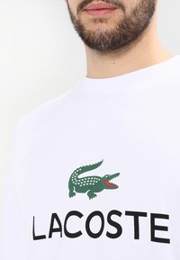 Biała bawełniana koszulka z logo zielonego krokodyla i czarnym napisem "LACOSTE" na przodzie. Klasyczny krój z okrągłym dekoltem.