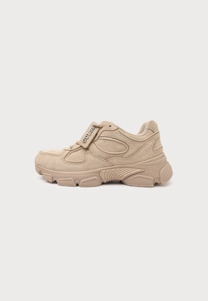 Baskets en daim beige avec une semelle épaisse, des coutures détaillées et une étiquette métallique avec le logo "GUESS" sur les lacets, vues de côté sur un fond uni.