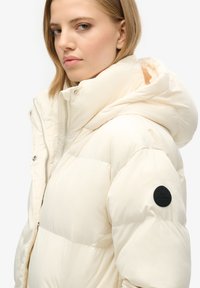 Creme farvet pufferjakke med aftagelig hætte, der har lynlås og en blød, quiltet tekstur. Sort logo-patch på venstre ærme.