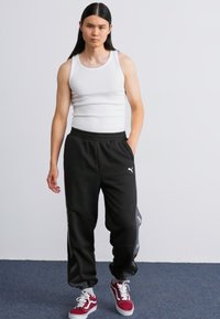 Fekete melegítőnadrág fehér Puma logóval, húzózsinórral és oldalsó zsebekkel, fehér ujjatlan toppal és piros sportcipőkkel párosítva.