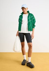 Grønn vindjakke med hvite detaljer, hvit t-skjorte, svarte sykkelshorts og svarte plattformklosser. Holder en teksturert hvit veske.