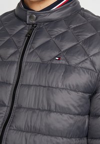 Grijze doorgestikte jas met een hoge kraag, een voorrits en contrasterende gestreepte bies. Heeft een klein logo-embleem op de borst.