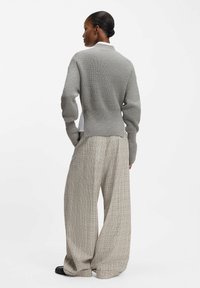 Woman wearing a gray knit sweater and beige wide-leg plaid trousers, standing with hands in pockets facing away.