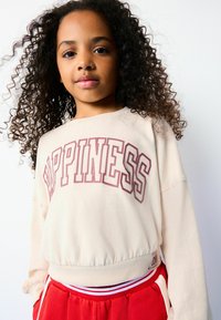 Beiger Sweatshirt mit "HAPPINESS" in kastanienbraun bedruckt, mit geripptem Saum und langen Ärmeln, kombiniert mit roten Jogginghosen mit weißen Akzenten.