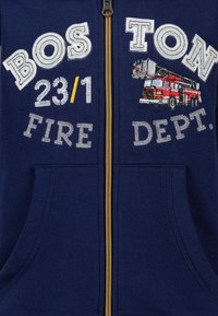Marine blauwe rits hoodie met geborduurde tekst "BOSTON 23/1 BRANDWEER" en een afbeelding van een rode brandweerwagen; bevat zijzakken en een gele ritssluiting.
