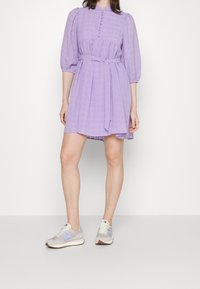 Lila Baumwollkleid aus strukturiertem Stoff, mit Puffärmeln, Knopfdetails und tailliertem Band. Kombiniert mit hellgrauen Sneakers mit blauen Akzenten.