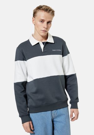 Joven con una camiseta de rugby gris oscuro y blanca con el texto "Simple is timeless", combinado con jeans azul claro, de pie sobre un fondo blanco.