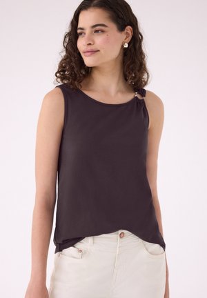 Jeune femme portant un haut sombre sans manches avec un détail en anneau sur l'épaule et un pantalon clair, regardant sur le côté.