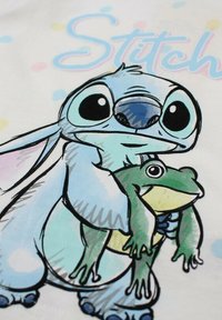 T-shirt en coton présentant un personnage de dessin animé aux grands yeux, fourrure bleue, et une grenouille. Le fond est orné de pois pastel et le texte "Stitch."
