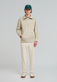 Beige zip-up sweatshirt with side pockets, paired with cream pants and beige sneakers. The model wears a green cap. Simple design, soft texture.