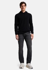 Schwarzer Hoodie mit Kordelzug, slim fit; kombiniert mit dunkelgrauen Jeans. Trägt weiße und graue Sneakers und steht vor einem schlichten Hintergrund.