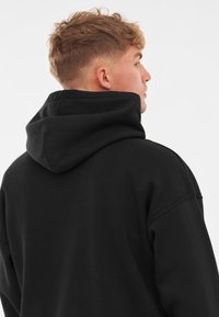 Sudadera negra con una capucha grande y estructurada, y un corte holgado. La tela parece suave, con una textura lisa y un mínimo detalle en las costuras.